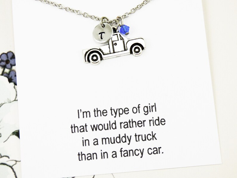 Op de afbeelding: Een zilveren ketting met een hanger in de vorm van een pick-up truck, een blauwe kristal en een zilveren schijf met de letter "T" erop. De ketting wordt geleverd met een kaart waarop staat: "Ik ben het type meisje dat liever in een modderige truck rijdt dan in een chique auto."