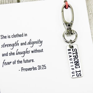 Puede incluir: Un llavero plateado con un dije que dice "Strong is Beautiful" y una tarjeta blanca con la cita "Ella está vestida de fuerza y dignidad y se ríe sin miedo al futuro. - Proverbios 31:25"