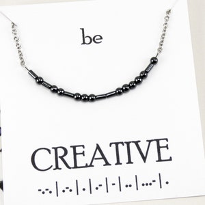 May include: A silver chain necklace with black beads that spells out "be creative" in braille. The necklace is on a white card with the word "be" at the top and the word "creative" in large letters below the necklace.