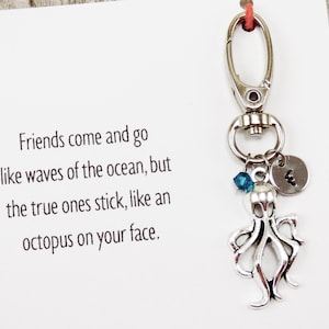May include: Silver octopus keychain with a blue crystal charm and a silver charm with the letter "E" on it. The keychain is attached to a card with the text "Friends come and go like waves of the ocean, but the true ones stick, like an octopus on your face."