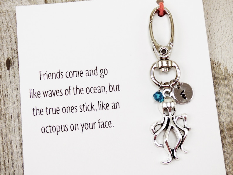 May include: Silver octopus keychain with a blue crystal charm and a silver charm with the letter "E" on it. The keychain is attached to a card with the text "Friends come and go like waves of the ocean, but the true ones stick, like an octopus on your face."