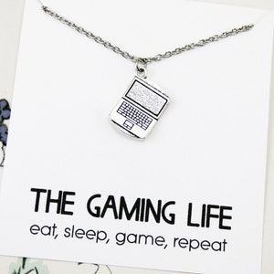 Może przedstawiać: Srebrny naszyjnik z zawieszką w kształcie laptopa. Zawieszka ma fakturowaną powierzchnię i czarny kontur. Naszyjnik jest dostarczany z kartą, na której napisano "The Gaming Life eat, sleep, game, repeat."