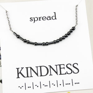 May include: A necklace with a chain and dark beads arranged to spell "KINDNESS" in Morse code. The word "spread" is above the necklace. The necklace is displayed on a white card.