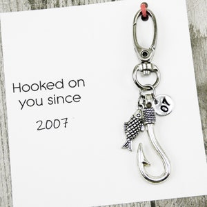 Puede incluir: Llavero de plata con un dije de pez, un dije de gancho y un dije con el número 07. Una tarjeta con el texto "Hooked on you since 2007" está en el fondo.