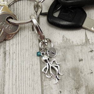 May include: Silver octopus keychain with a blue bead and a silver charm with the letter "K" on it. The keychain is attached to a silver key ring with a set of keys.