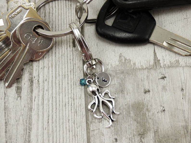 May include: Silver octopus keychain with a blue bead and a silver charm with the letter "K" on it. The keychain is attached to a silver key ring with a set of keys.