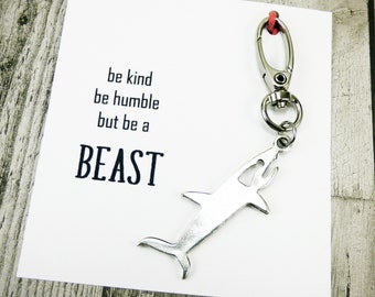 Op de afbeelding: Een witte kaart met de tekst "be kind be humble but be a BEAST" en een zilveren haai sleutelhanger eraan bevestigd.