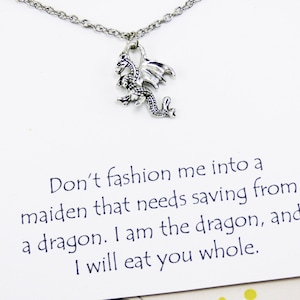May include: A silver-colored dragon pendant necklace. The dragon charm is detailed with wings and scales, suspended from a delicate chain. The card behind the necklace reads, "Don't fashion me into a maiden that needs saving from a dragon. I am the dragon, and I will eat you whole."