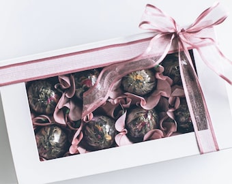 May include: A white gift box with a pink ribbon and bow. The box is filled with loose leaf tea balls, each wrapped in pink tissue paper.