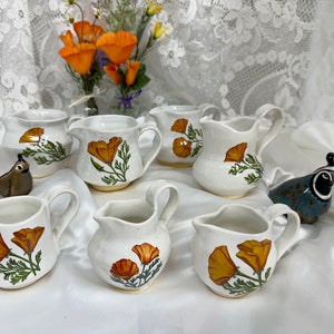 以下が含まれることがあります： オレンジ色のカリフォルニアポピーの花柄が描かれた白いセラミック製のクリーマーピッチャー7個セット。ピッチャーは白い表面に並べられ、小さな茶色のウズラ像2体と青と茶色のウズラ像1体が置かれています。