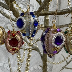 Puede incluir: Adornos decorativos en forma de huevo, en varios colores, con ganchos de alambre dorado. Los adornos presentan varios colores, incluyendo oro, rojo, azul, púrpura y verde, y están adornados con gemas y perlas falsas. Cuelgan de una rama decorativa.