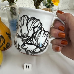 May include: White ceramic mug with a black floral design. The mug has a curved shape and a handle. The floral design is a bold, stylized poppy. The mug is held by a person with orange nail polish. A small white square with the letter "H" is in the image.
