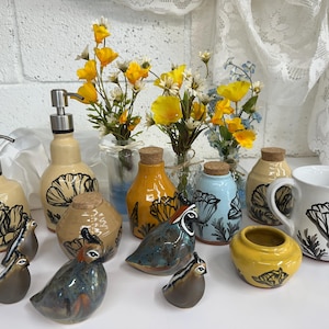 May include: A collection of ceramic items, including soap dispensers, bottles, a mug, and small bird figurines. The items are in various colors, including beige, yellow, and blue, with black floral designs. Small vases with yellow flowers are also present.