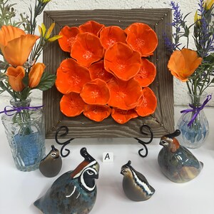 Puede incluir: Una exhibición con figuras de cerámica de codornices en varios colores, incluyendo azul, marrón y patrones moteados. Una pieza de arte mural enmarcada en madera con acentos de flores de color naranja brillante está en el fondo, junto con jarrones de flores.