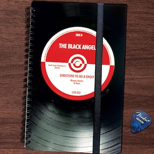 Könnte beinhalten: Schwarzes Vinyl-Notizbuch mit einem roten und weißen Schallplattenlabel, auf dem "The Black Angel" und "Directions to See a Ghost" steht. Das Label enthält auch den Text "Mission District, 18 Years, LITA 033".