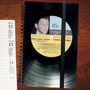 Könnte beinhalten: Ein schwarz-weißes Spiralnotizbuch mit einem Vinyl-Plattencover, das ein Foto von Frank Sinatra und den Text "Francis Albert Sinatra" und "Antonio Carlos Jobim" zeigt. Das Plattenlabel ist gelb mit schwarzem Text. Das Notizbuch hat ein schwarzes Gummiband.