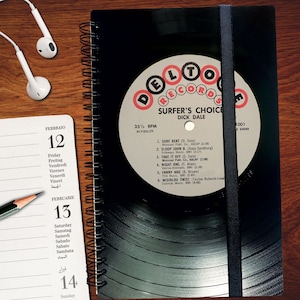 Könnte beinhalten: Ein schwarz-weißes Notizbuch mit Spiralbindung. Der Einband zeigt ein Vintage-Schallplattenlabel mit dem Text "Delto Records Surfer's Choice Dick Dale" und einer Liste von Songtiteln. Das Notizbuch ist eine großartige Möglichkeit, Ihre Gedanken, Ideen und Erinnerungen festzuhalten.