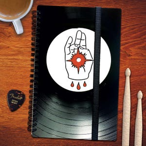 Könnte beinhalten: Ein schwarzes Spiralnotizbuch mit einem weißen Kreis auf der Vorderseite. Der Kreis zeigt eine Schwarzweißillustration einer Hand mit einem roten Stern in der Handfläche. Das Notizbuch hat ein schwarzes Schallplatten-Design auf der Rückseite.