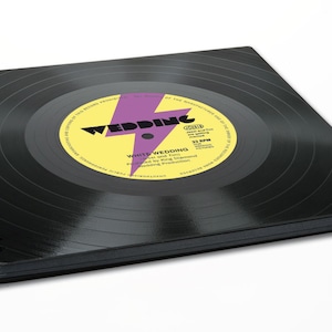 Peut inclure: Carnet à spirales noir avec un design de disque vinyle. L'étiquette présente un fond jaune avec un éclair violet et le texte "WEDDING" en noir. L'étiquette comprend également le texte "WHITE WEDDING" et "33 RPM".