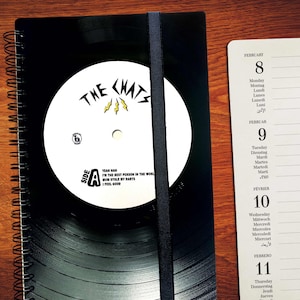 Könnte beinhalten: Ein schwarzes Spiralnotizbuch mit einem schwarzen Vinyl-Platten-Design auf dem Cover. Die Platte hat ein weißes Etikett mit dem Text "THE CHATS" und "SIDE A" in Gelb. Der Text auf dem Etikett lautet "YEAH NAN I'M THE BEST PERSON IN THE WORLD MUM STOLE MY DARTS I FEEL GOOD". Das Notizbuch liegt neben einer Kalenderseite mit dem Monat Februar.