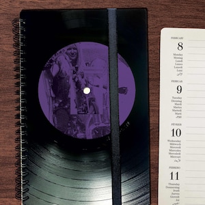 Könnte beinhalten: Ein schwarzes Spiralblock mit einer violetten Schallplatte auf dem Cover. Die Schallplatte zeigt ein Schwarzweißbild einer Person auf einem Motorrad. Das Notizbuch hat einen schwarzen Gummibandverschluss.