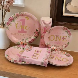 May include: Pink and gold floral themed party supplies for a first birthday. The set includes plates, napkins, and cups. The plates feature a floral design with the number one and the words "Happy Birthday". The napkins have a similar design with the word "One". The cups are pink with a white rim.