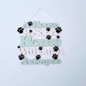 Op de afbeelding: Een houten bord met de tekst "A House Is Not A Home Without A Jackapoo" in zwart-witte letters op een groene en witte achtergrond. Het bord is versierd met zwarte poot afdrukken.