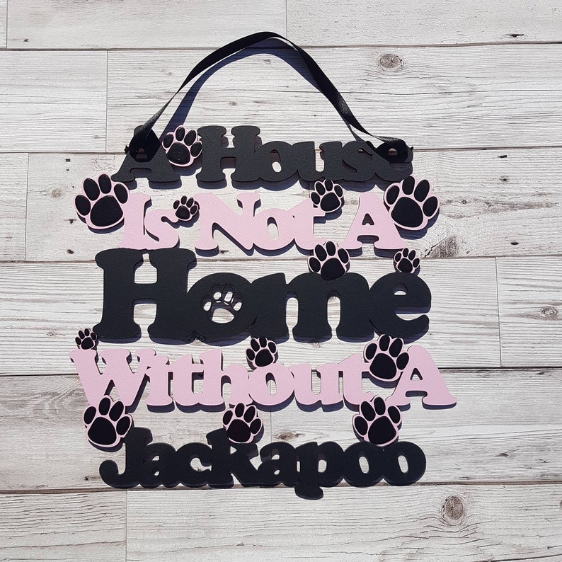 Op de afbeelding: Een zwart en roze houten bord met de tekst "A House Is Not A Home Without A Jackapoo". Het bord is versierd met poot afdrukken.