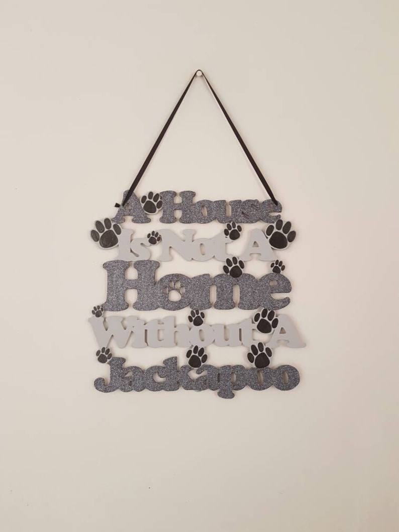 Op de afbeelding: Een wit en grijs houten bord met zwarte poot afdrukken en de tekst "A House Is Not A Home Without A Jackapoo".