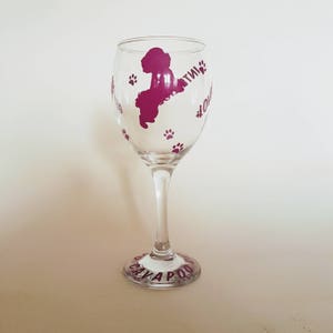 May include: A clear glass wine glass with a pink silhouette of a dog and paw prints. The text "Cavapoo" is written on the bottom of the glass.