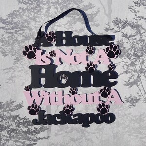 Op de afbeelding: Een zwart en roze houten bord met de tekst "A House Is Not A Home Without A Jackapoo" en hondenpootafdrukken.