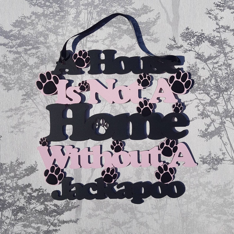 Op de afbeelding: Een zwart en roze houten bord met de tekst "A House Is Not A Home Without A Jackapoo" en hondenpootafdrukken.