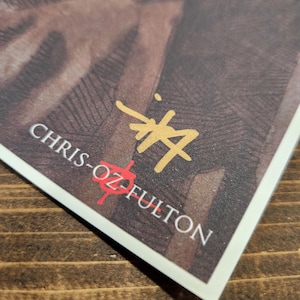 May include: A close-up of a signature by Chris-Oz Fulton, written in gold ink on a white background. The signature is on a brown textured surface.