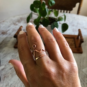 May include: A copper-colored wire ring with an abstract design. The ring features a wavy line and a loop, creating a unique and artistic look. The ring is worn on a finger, showcasing its delicate craftsmanship and minimalist style.