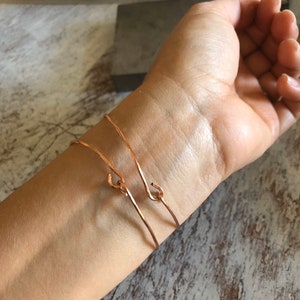May include: Two thin rose gold bracelets with a simple hook closure. The bracelets are worn on a person's wrist.
