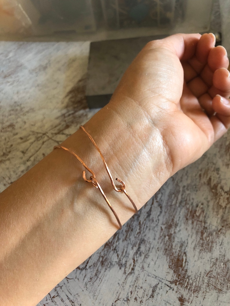 May include: Two thin rose gold bracelets with a simple hook closure. The bracelets are worn on a person's wrist.