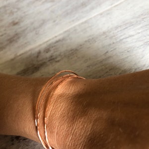 May include: Three thin, hammered copper bangles stacked on a wrist. The bangles are a warm, reddish-brown color.