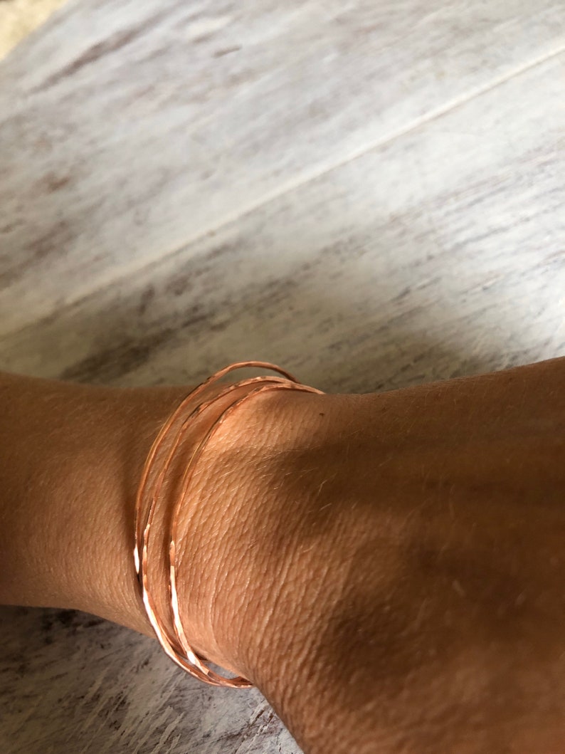 May include: Three thin, hammered copper bangles stacked on a wrist. The bangles are a warm, reddish-brown color.
