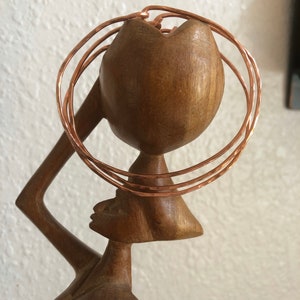 May include: A wooden sculpture of a woman with a copper wire headpiece. The headpiece is made of multiple rings of copper wire, creating a layered effect.
