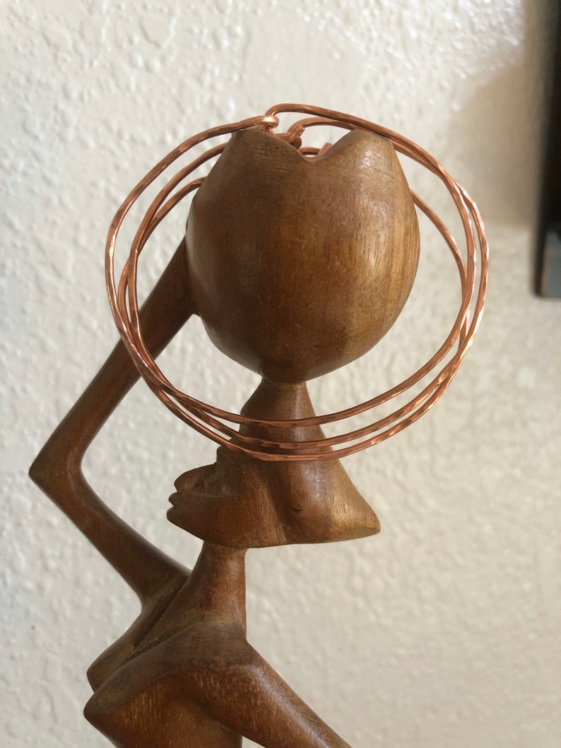 May include: A wooden sculpture of a woman with a copper wire headpiece. The headpiece is made of multiple rings of copper wire, creating a layered effect.