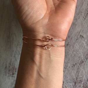 May include: Two thin rose gold bracelets with a knot detail on the wrist of a person.