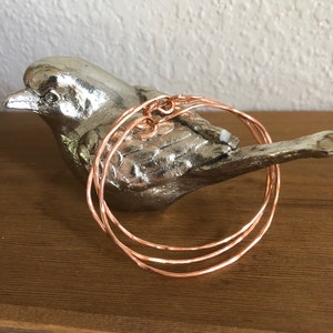 May include: Three thin, rose gold colored, hammered metal bangles stacked on top of each other.