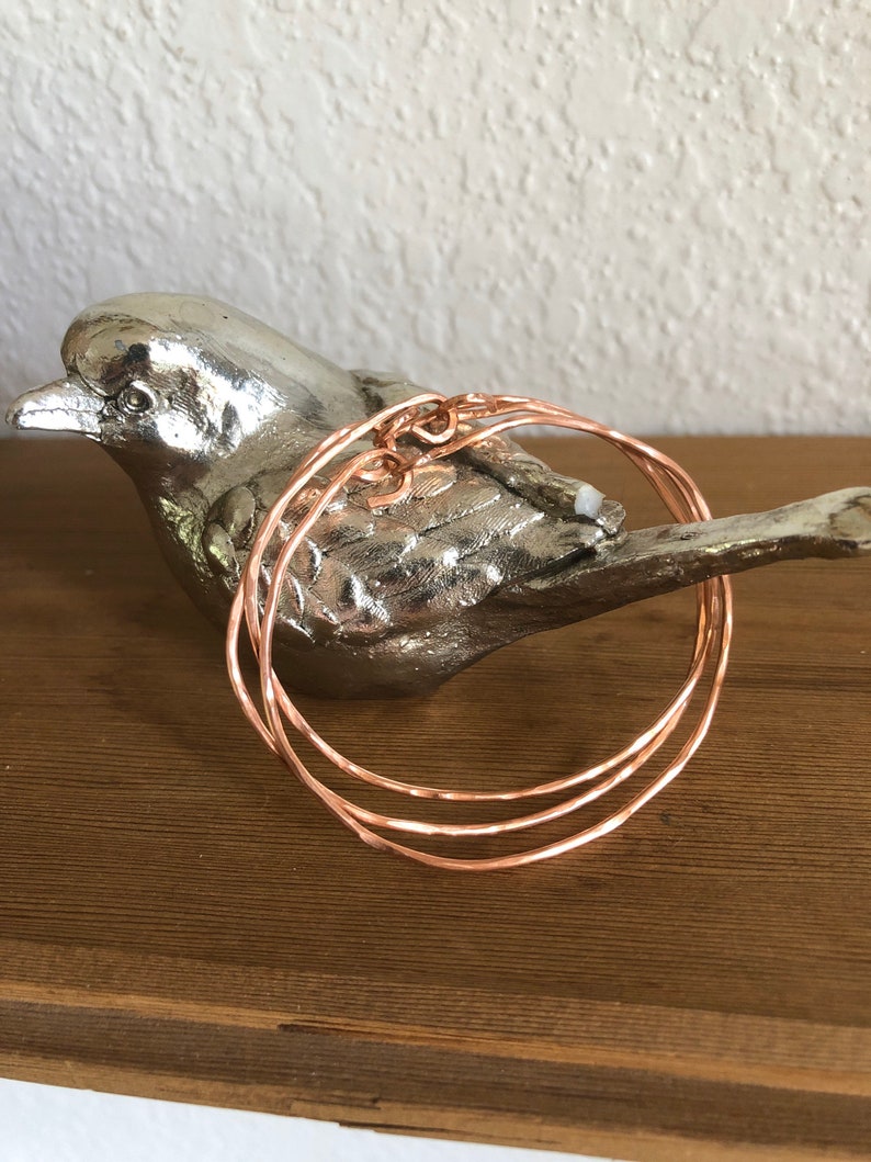 May include: Three thin, rose gold colored, hammered metal bangles stacked on top of each other.