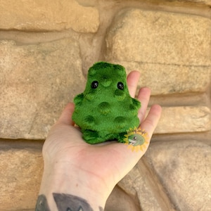Könnte beinhalten: Ein kleiner, grüner Plüschfrosch mit schwarzen Augen und einer strukturierten Oberfläche. Der Frosch wird in einer Hand vor einem Hintergrund aus beigem Stein gehalten. Der Frosch ist ein handgefertigter Artikel.