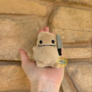 Puede incluir: Un juguete de peluche beige con forma de patata con una cara sonriente y un cuchillo pequeño. El juguete se sostiene en una mano sobre un fondo de piedra texturizada. El juguete tiene ojos negros y boca negra.