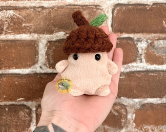 Puede incluir: Un pequeño peluche con forma de bellota con cuerpo beige claro, un sombrero de ganchillo marrón oscuro y una pequeña hoja verde. El juguete tiene dos ojos de cuentas negras y un logotipo con una rana y el texto Stitching Sunlight.