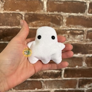 Puede incluir: Un pequeño juguete de peluche de fantasma blanco con una textura suave y difusa y dos ojos redondos de plástico negro. El fantasma tiene una cabeza redondeada y dos brazos pequeños extendidos, con un borde inferior ondulado y festoneado.