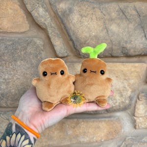 May include: Two brown plush toys shaped like sprouts, with black eyes and small limbs. One has a green sprout on top. They are held in a hand with an orange bracelet and a sunflower tattoo.