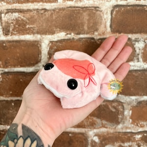 May include: A small, light pink plush frog toy with large black plastic eyes and a soft, fuzzy texture. The frog features a darker pink patch on its head with a red embroidered bow detail. A small circular logo with the text Stitching Sunlight is visible.