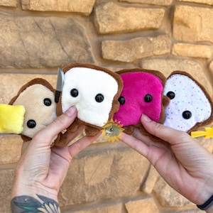 May include: Four plush bread slices with brown crusts and black bead eyes. The first has a yellow butter square, the second a small silver knife, the third a bright pink topping, and the fourth a white surface with colorful sprinkles and a yellow star.